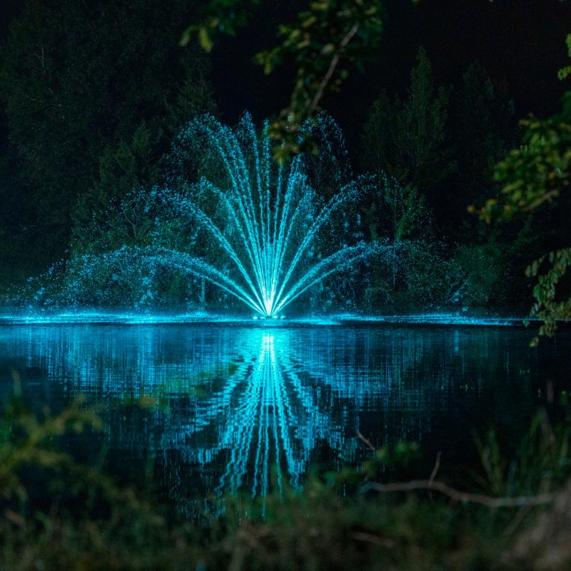 Kasco Mighty Oak Fountain Masterpiece Bundle illuminated at night with teal lighting in wooded pond setting
