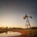 Outdoor Water Solutions Super Premier 3-Leg Windmill Pond Aerator installed near a pond shoreline with farm buildings in the background at sunset